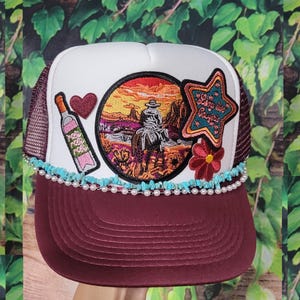 May include: A burgundy and white trucker hat with a mesh back. The hat features a circular patch with a desert scene and a rider on horseback, a star-shaped patch with text, and a bottle of rosé. Embellished with turquoise stones and pearls.