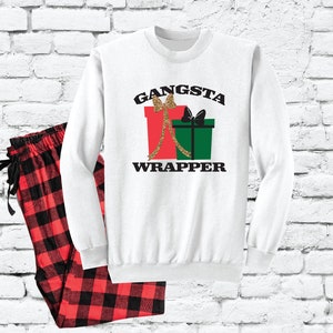 Puede incluir: Sudadera blanca de manga larga con el texto "Gangsta Wrapper" y un gráfico de dos regalos envueltos con cinta dorada y lazos. Pantalón de pijama a juego de cuadros rojos y negros.