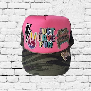 May include: Pink trucker hat with a camouflage brim. The hat has several patches, including a peace sign, a lightning bolt, a jeep, and the words "Just Have Fun".