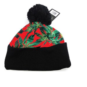 Pode incluir: Boné vermelho com um pompom preto e um padrão de folhas verdes. O boné tem uma aba preta.