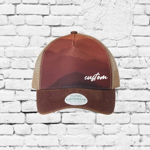 May include: A brown and tan trucker hat with a sunset mountain scene and the word "custom" in white script. The hat has a brown leather bill and a brown mesh back. The hat has a green and white label that says "Old Favorite Structured Legacy".