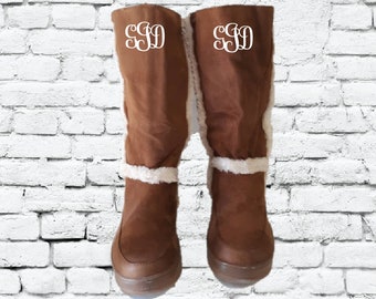 Op de afbeelding: Een paar bruine suède laarzen met witte nep bont trim en een wit monogram "S.S.D." op elke laars.