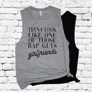 May include: Gray sleeveless muscle tank top with black text that reads "Tryna look like one of those rap guys girlfriends".