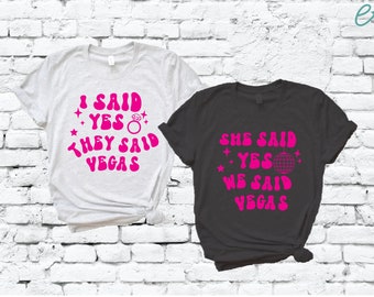 Puede incluir: Dos camisetas con un diseño de fuente retro rosa. La camiseta blanca dice "Dije que sí, ellos dijeron Las Vegas" con un anillo y estrellas. La camiseta negra dice "Ella dijo que sí, nosotros dijimos Las Vegas" con una bola de discoteca y estrellas.