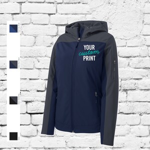 May include: A navy blue and grey softshell jacket with a zip fastening and a hood. The jacket has a contrasting colour panel on the chest and sleeves. The text "YOUR custom PRINT" is embroidered on the chest in teal.