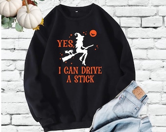 May include: Black sweatshirt with a white graphic of a witch riding a broomstick with a black cat. The text "YES, I CAN DRIVE A STICK" is printed in orange below the graphic.