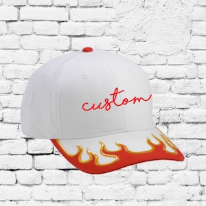 Może przedstawiać: Biała czapka bejsbolówka z haftem "custom" w kolorze czerwonym z przodu. Daszek czapki jest czerwony z żółtymi płomieniami.