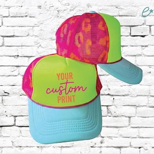 May include: Two trucker hats. One hat has a neon green front panel with the text "YOUR custom PRINT" in pink script. The other hat has a pink mesh top with an orange pattern, a neon green brim, and a light blue bill.
