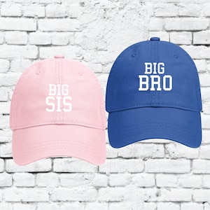 Puede incluir: Dos gorras de béisbol, una rosa y otra azul, con las palabras "BIG SIS" y "BIG BRO" bordadas en blanco en la parte delantera.
