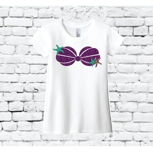 Puede incluir: Camiseta blanca con un diseño de concha de sirena de purpurina morada y estrellas de mar de purpurina verde y marrón.