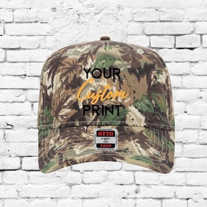 May include: A camouflage print baseball cap with the text "YOUR Custom PRINT" in a script font. The cap has a classic fit and a curved brim. A small tag on the side reads "OTTO Classic Fit Camo".