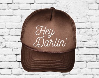 May include: A brown trucker hat with a white mesh back. The hat has white cursive text that says "Hey Darlin'" on the front.
