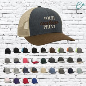 May include: A collection of trucker hats in various colors, including black, gray, and blue. The main hat features a dark gray front panel, a brown brim, and a tan mesh back. The text "YOUR custom PRINT" is embroidered on the front.