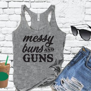 Puede incluir: Camiseta de tirantes gris con el texto "messy buns and guns" en letras negras.