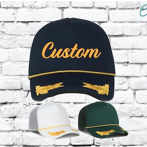 Könnte beinhalten: Marineblaue Trucker-Mütze mit dem Wort "Custom" in goldener Schrift. Die Mütze hat goldene Verzierungen und goldene Stickereiakzente. Zwei weitere Trucker-Mützen sind abgebildet, eine weiße und eine grüne, beide mit goldenen Akzenten.