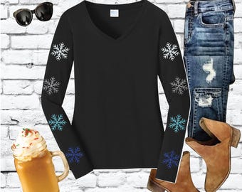 May include: A black long-sleeve V-neck shirt with snowflake designs on the sleeves, paired with distressed blue jeans and brown suede ankle boots. The image also includes a mug of a beverage with whipped cream and sunglasses.
