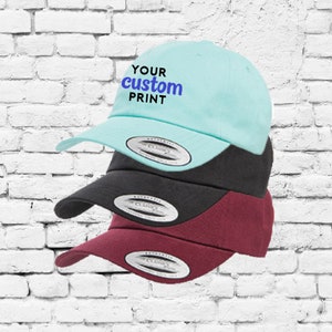 May include: Three baseball caps stacked on top of each other. The top cap is light blue with the text "YOUR custom PRINT" embroidered on the front. The middle cap is black and the bottom cap is burgundy.