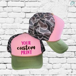 Puede incluir: Dos gorras de camionero de camuflaje rosa y verde. Una gorra tiene el texto "YOUR custom PRINT" en la parte delantera.