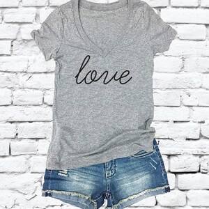 Può includere: Una maglietta grigia a collo a V con la parola "love" scritta in corsivo sul davanti. La maglietta è abbinata a pantaloncini di jeans.