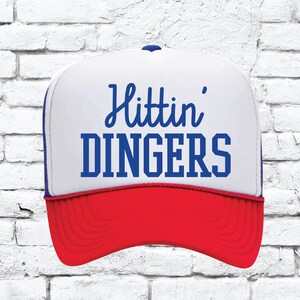 May include: A white and red trucker hat with a blue mesh back. The front of the hat has the text "Hitting Dingers" in blue.