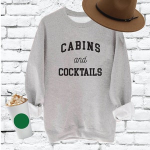 Puede incluir: Una sudadera gris con el texto "CABINS and COCKTAILS" impreso en negro. La sudadera está colocada sobre un fondo de ladrillos blancos.