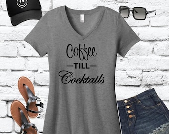 May include: A grey V-neck t-shirt with the text "Coffee - Till - Cocktails" in black script. The image also includes a black baseball cap with a smiley face, denim shorts, sunglasses, and brown sandals.