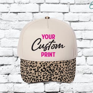May include: A beige baseball cap with a leopard print brim. The cap features the words "YOUR Custom PRINT" in black and pink lettering. The hat is set against a white brick wall.