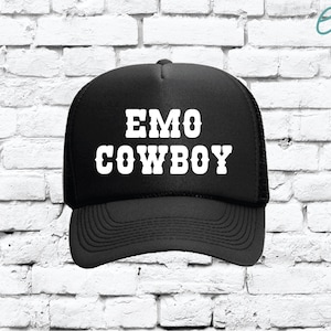 以下が含まれることがあります： 前面に白い文字で「EMO COWBOY」と書かれた黒いトラッカーハット。帽子は湾曲したつばとメッシュの背面を備えています。帽子は白いレンガの壁を背景にしています。