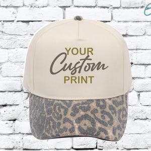May include: A beige baseball cap with a leopard print brim. The cap features the text "YOUR Custom PRINT" in a dark olive green colour. The cap is set against a white brick wall background.