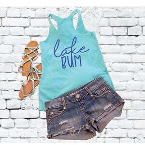 Puede incluir: Un top de tirantes azul claro con el texto "lake bum" en letras azules. El top se combina con pantalones cortos de mezclilla y sandalias.