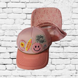 Pode incluir: Boné de caminhoneiro rosa com vários patches, incluindo um sinal de paz, uma prancha de surf com o texto "beach vibes", um rosto sorridente e um cupcake. O boné também tem o texto "Take it easy" na parte da frente.