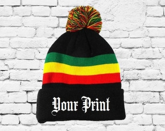 May include: A black knit beanie featuring horizontal stripes in green, yellow, and red above the cuff. The cuff displays the white text "Your Print" in a gothic font. A multicolored pom-pom in red, yellow, and green sits on the top of the hat.