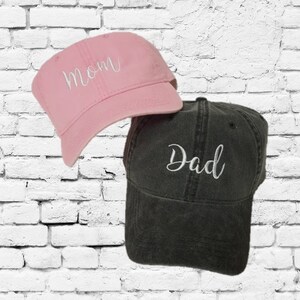 May include: Two baseball caps, one pink with the word "Mom" embroidered in white, and one black with the word "Dad" embroidered in white.