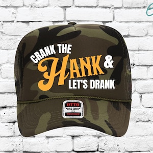 May include: A camouflage trucker hat with the text "CRANK THE HANK & LET'S DRANK" in white and orange lettering. The hat has a mesh back and a curved brim. The hat also has a small tag that says "OTTO WORLD FAMOUS FOAM FRONT".