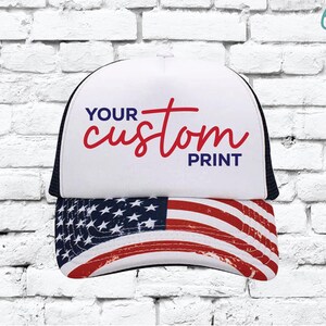 May include: A white trucker hat with a black mesh back and a curved brim featuring the American flag. The front panel displays the text "YOUR custom PRINT" in red and blue.