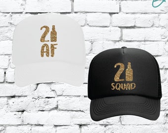 Puede incluir: Dos gorras de camionero con texto de purpurina dorada. Una gorra es blanca con el texto "21 AF" y la otra es negra con el texto "21 Squad".
