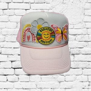 Puede incluir: Gorra de camionero rosa con una parte trasera de malla blanca. La gorra tiene varios parches bordados, incluyendo un arcoíris, una flor, un sol con el texto "Radiated Positivity" y una mariposa.