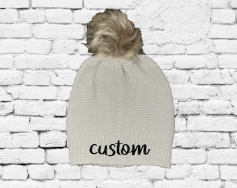 May include: A white knit beanie with a faux fur pom pom and the word "custom" embroidered on the front.