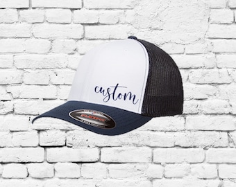 May include: White and black trucker hat with a navy blue brim. The hat has a mesh back and the front features the word "custom" in blue cursive writing.
