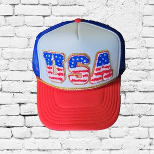 May include: A white and red trucker hat with a blue mesh back. The front of the hat features a large embroidered patch that says "USA" in white letters with red and blue stars. The letters are outlined in gold.