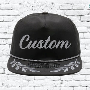 Puede incluir: Una gorra de béisbol negra y gris bordada a medida con un ribete de cuerda gris y un diseño de hojas bordadas grises en la visera. La gorra dice "Custom" en una fuente cursiva gris.
