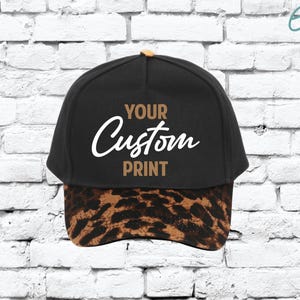 May include: Black baseball cap with a leopard print brim and a tan button on top. The cap features the text "YOUR Custom PRINT" in white and tan lettering.