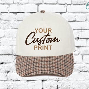 May include: A cream-colored baseball cap with a brown and tan plaid brim. The cap features the text "YOUR Custom PRINT" in brown script and block letters. The cap has a rounded top and a small button.