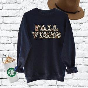 Pode incluir: Uma camisa de frio azul marinho com o texto "FALL VIBES" em letras douradas com um padrão de leopardo. A camisa de frio está em um fundo de tijolos brancos.