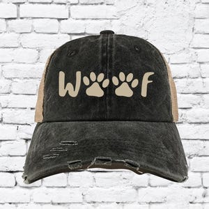 Op de afbeelding: Verweerde zwarte baseballcap met een bruin mesh achterkant. De voorkant toont het woord "Woof" in een crèmekleurig lettertype, met pootafdrukken in plaats van de "o" letters. De cap heeft een vintage uitstraling.
