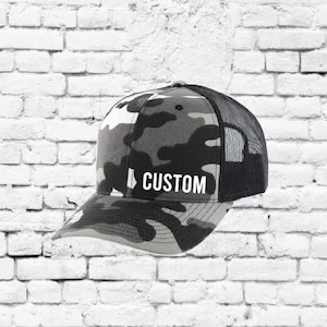 May include: A black and gray camouflage trucker hat with a black mesh back. The hat has the word "CUSTOM" embroidered in white on the front.