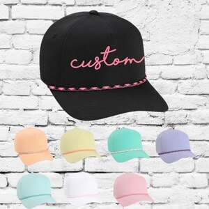 Puede incluir: Una gorra de béisbol negra con un ribete de cuerda rosa y la palabra "custom" bordada en rosa en la parte delantera. La gorra se muestra con varias otras gorras de béisbol en varios colores pastel.