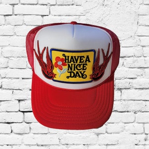 Puede incluir: Una gorra de camionero roja y blanca con un parche amarillo que dice "Have a Nice Day" con una flor y dos pájaros negros.