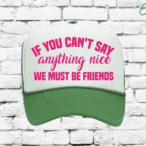 May include: A green and white trucker hat with the text "IF YOU CAN'T SAY anything nice WE MUST BE FRIENDS" in pink. The hat has a white front panel and a green brim and mesh back.