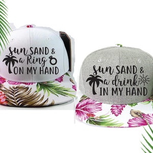 May include: Two baseball caps with a tropical print on the underside of the brim. The front of the caps have black text that reads "Sun Sand & a Ring on my hand" and "Sun Sand & a drink in my hand".